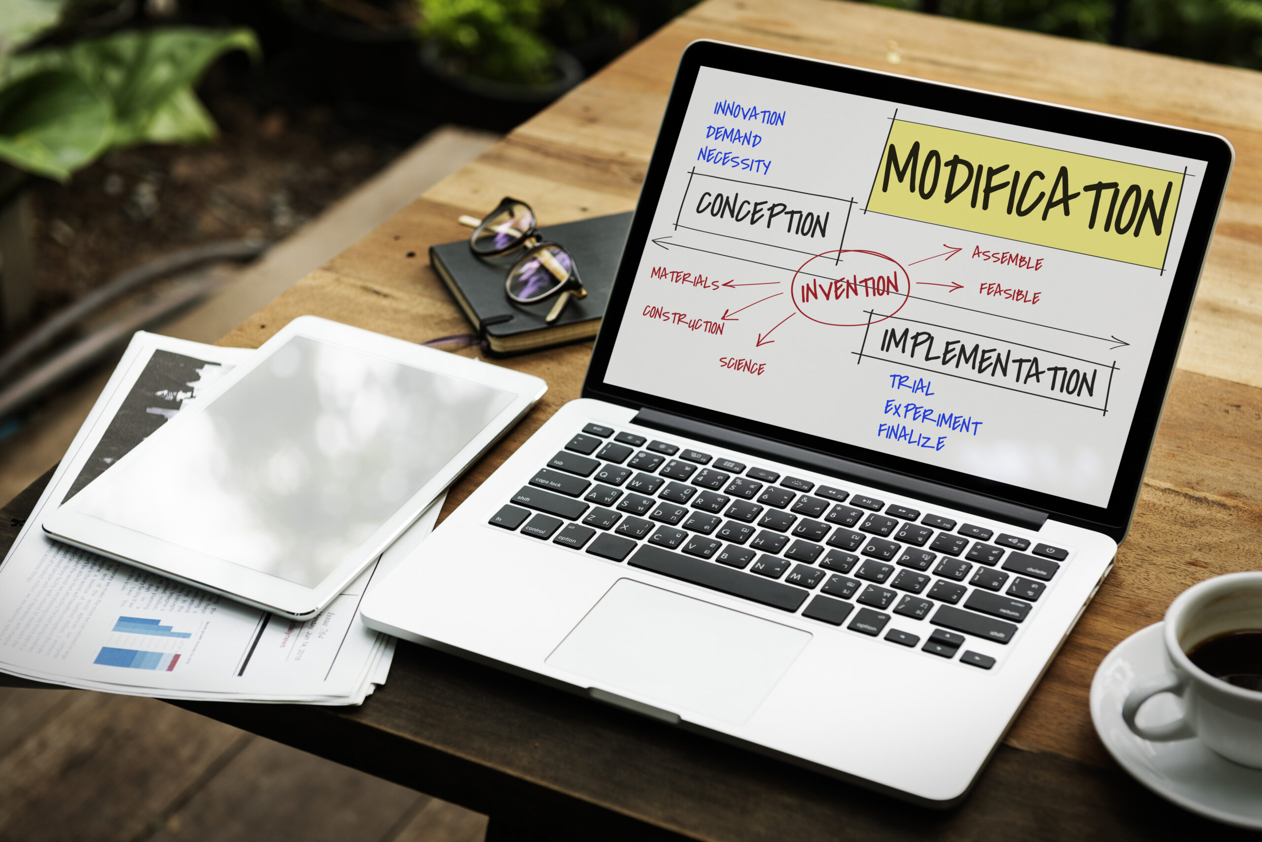 Laptop z wyświetlonym diagramem procesu innowacji – etapy: koncepcja, wdrożenie, modyfikacja; obok tablet, notatki i filiżanka kawy na drewnianym stole.